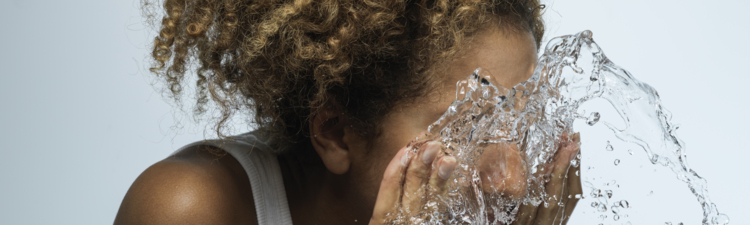 Young woman with dark skin and curly hair splashing face with fresh water against blue background