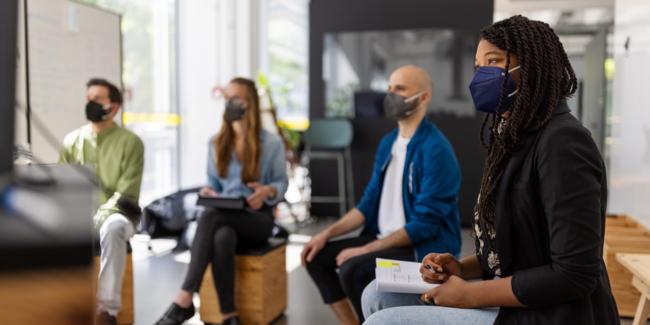 Multi-ethnic business people wearing face mask looking at television screen during online meeting. Men and women sitting at social distance during a video conference meeting in hybrid office.