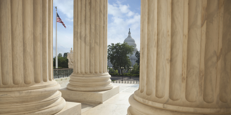 Close up of the columns  of the Supreme Court building with an American flag and the US Capitol in the background