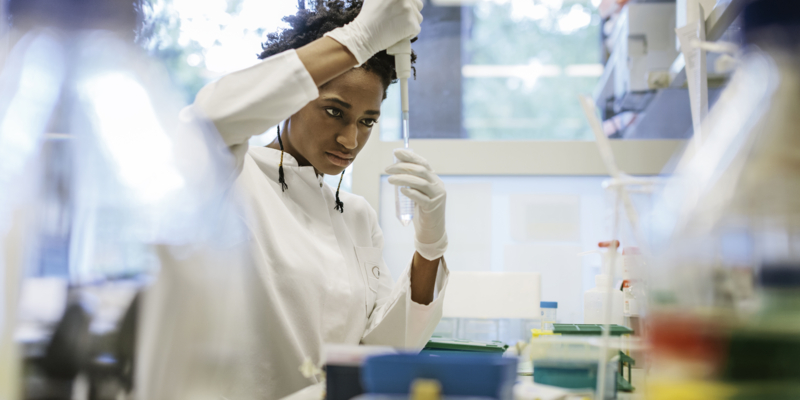 A black female scientist is pipetting in a laboratory. She's holding a pipette in one hand and a small flask in the other one, while wearing white gloves.