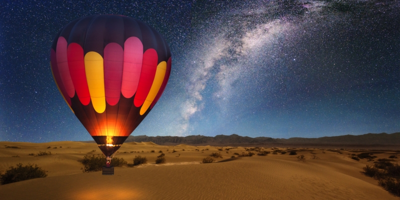 A majestic hot air balloon soars under the stars of the Milky Way, over the desert - Mesquite Dunes of Death Valley National Park. Moonlight provides luminosity showing the patterns and shapes of the desert landscape.